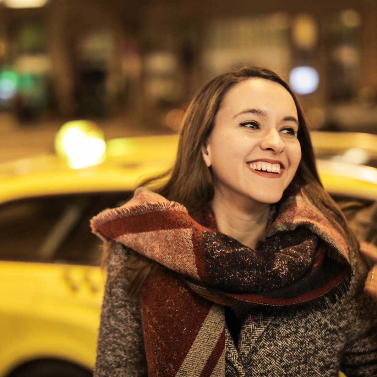 A smiling woman in a scarf on a city street at night, a yellow taxi behind her.